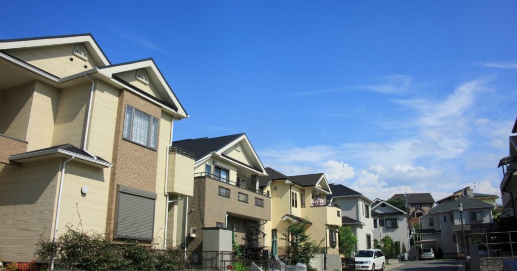 A residential neighborhood featuring a row of modern, multi-story homes with light-colored facades and sloped roofs. The houses are lined with small gardens and driveways, with a bright blue sky and scattered white clouds in the background, creating a calm and inviting atmosphere.